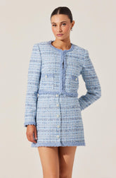 A woman is wearing a light blue, textured, tweed-style two-piece suit. The jacket has a round neckline, long sleeves, and is decorated with two rows of large, white buttons on the front. The skirt is a matching light blue, textured, tweed-style, high-waisted mini skirt with a row of white buttons down the front. The fabric appears to be woven with subtle multicolored threads, giving it a classic, elegant look.